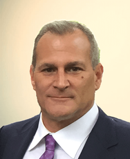 A man with short hair wearing a navy blue suit, white collared shirt, and purple tie, posed against a plain light wall.