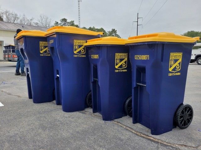 Four blue trash bins with yellow lids, lined up in an outdoor parking area near a person in the background.