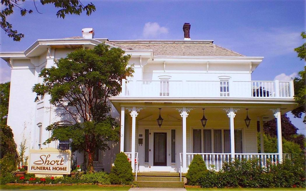 White Victorian-style house with a porch, trees, and a sign in front under a blue sky