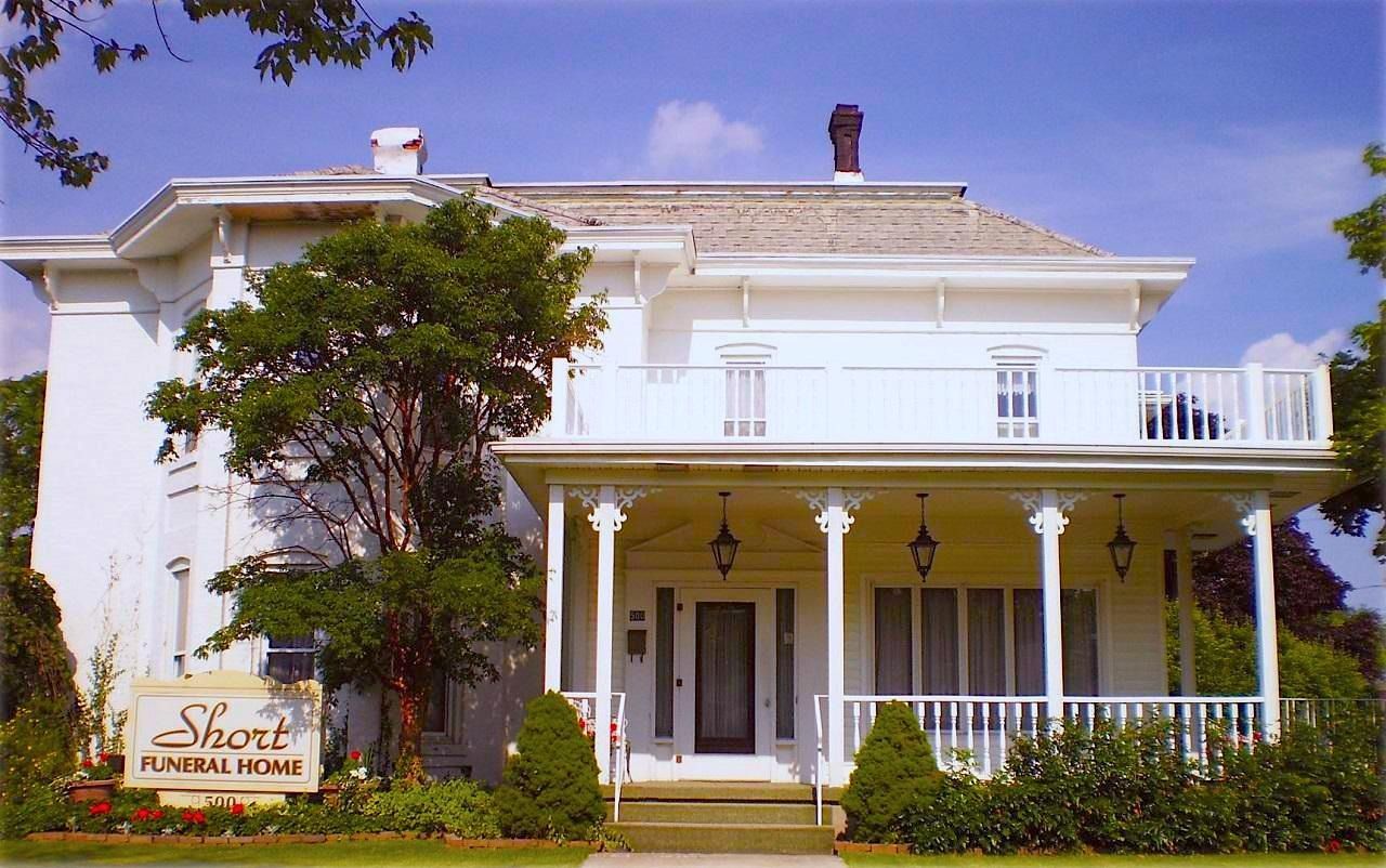 White Victorian-style house with a porch, trees, and a sign in front under a blue sky