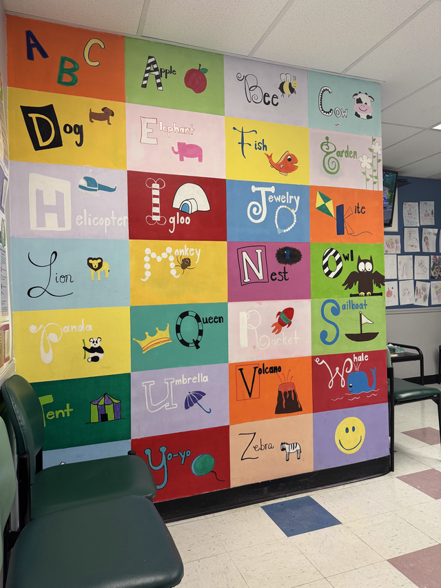 Colorful alphabet mural made of decorated square tiles on a hallway wall beside green chairs.