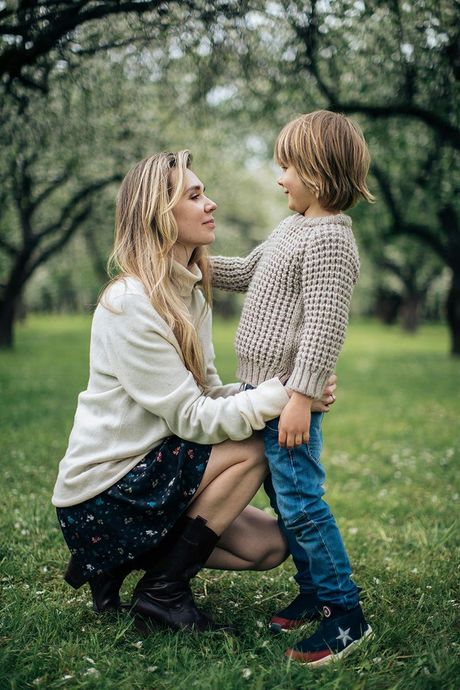 Woman kneels on grass, smiling at a child in a knit sweater and jeans in a leafy park.