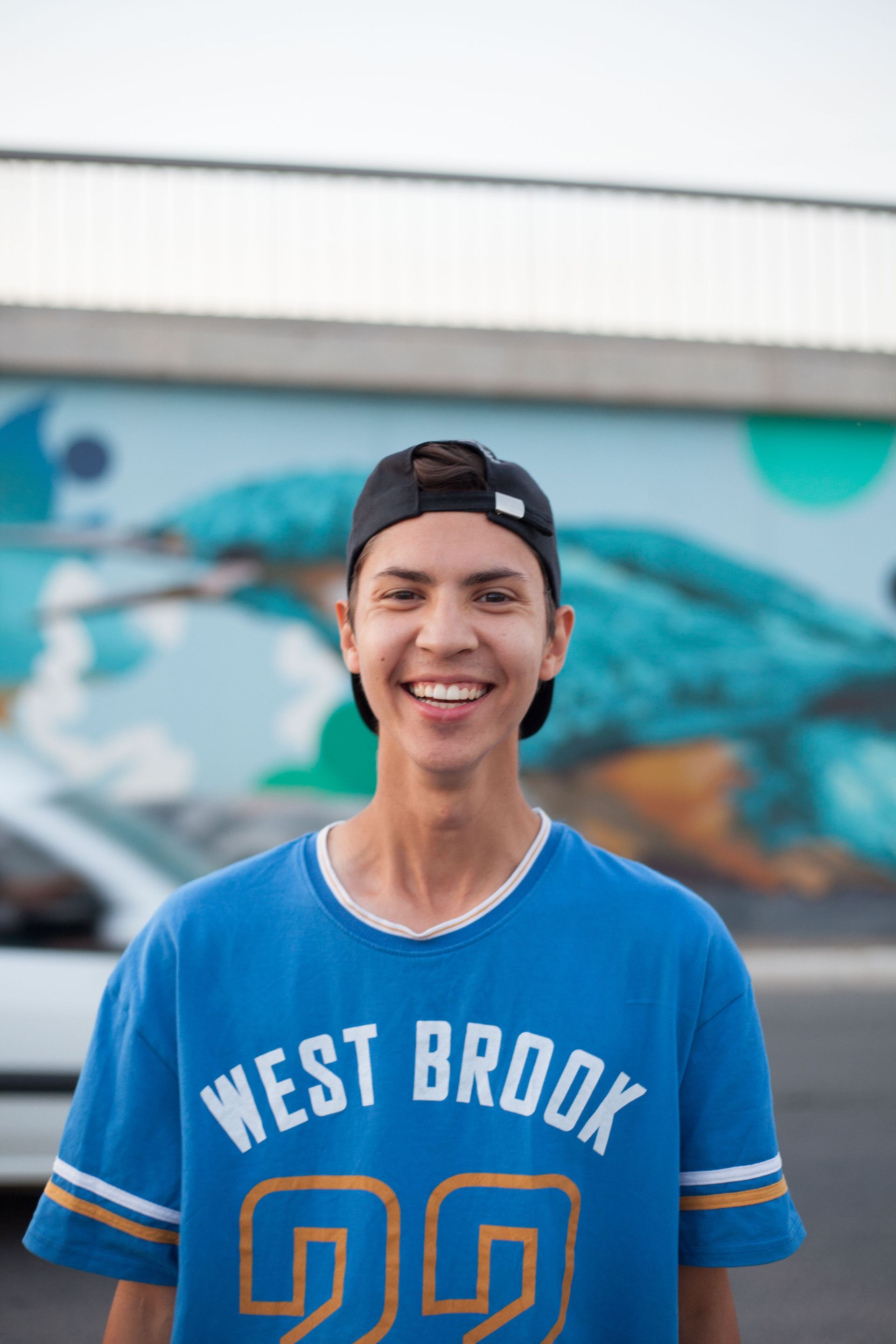 Smiling person in a blue WEST BROOK jersey and backward cap standing outdoors in front of a mural