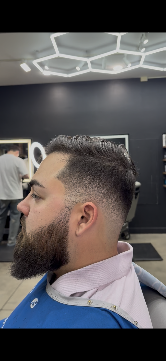Man with beard getting a haircut in a barbershop. He has a faded haircut and a white barber's neck protector.