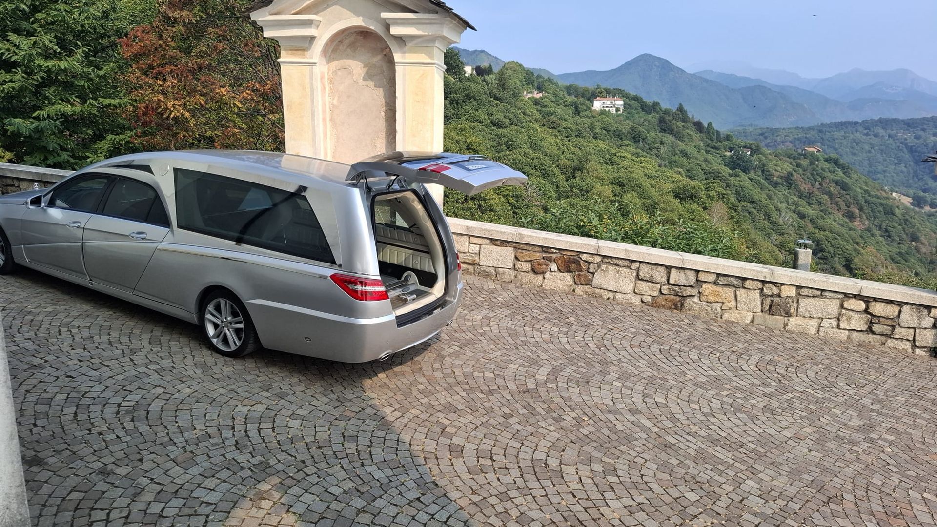Un minivan argentato parcheggiato su un punto panoramico lastricato, con montagne e vallate sullo sfondo.