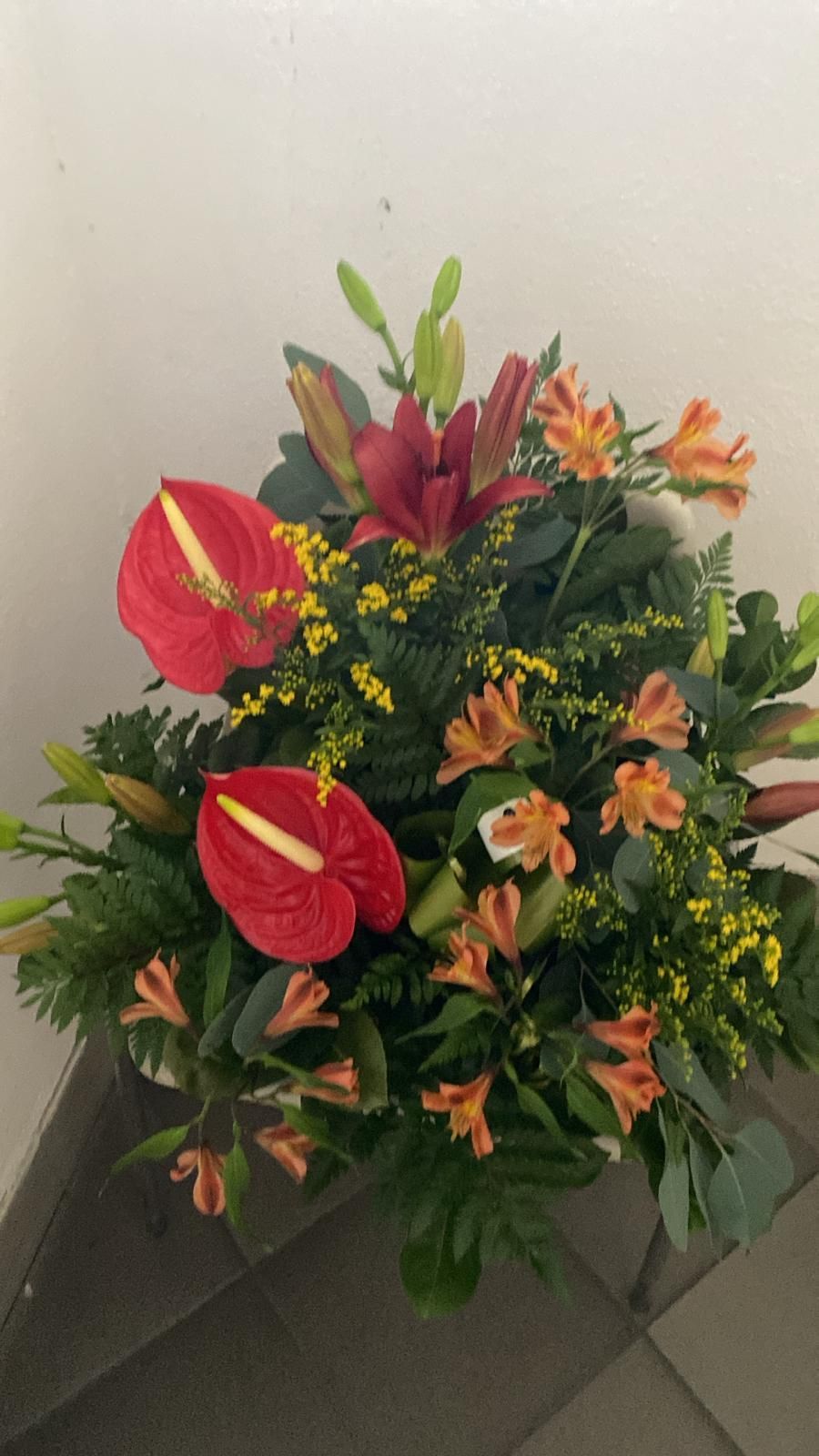 Bouquet con anthurium rossi, gigli arancioni e fiori gialli su foglie verde scuro.