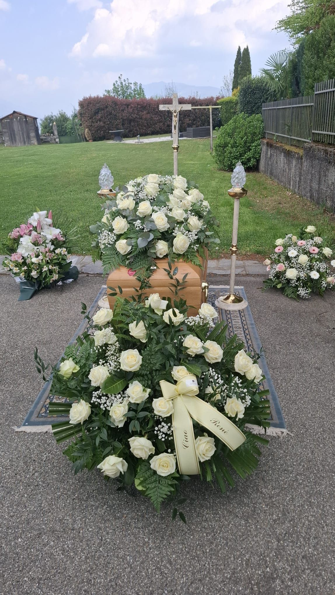 Corone e bouquet funebri floreali disposti su un sentiero di ghiaia in un cimitero-giardino.