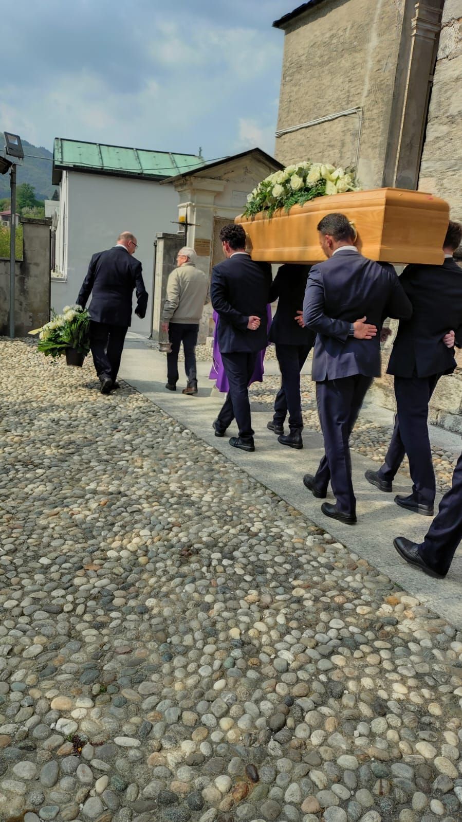 I portatori di bara trasportano una bara di legno leggero attraverso un cortile lastricato, accanto a edifici in pietra.