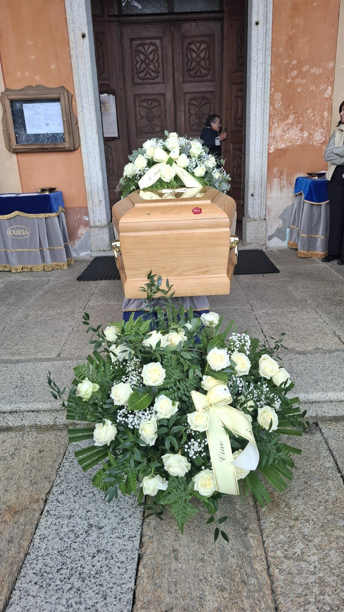 Corone funebri di fiori bianchi circondano una bara di legno chiaro all'ingresso di un edificio.