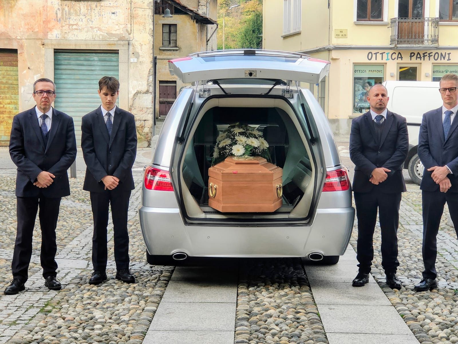 Uomini in abiti neri sono in piedi accanto a un carro funebre aperto che trasporta una bara su una strada acciottolata.