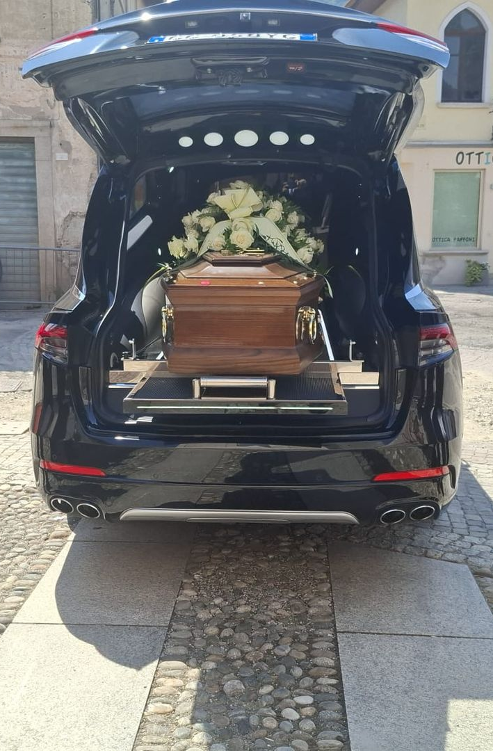 Carro funebre aperto con bara di legno e fiori bianchi durante un corteo funebre.