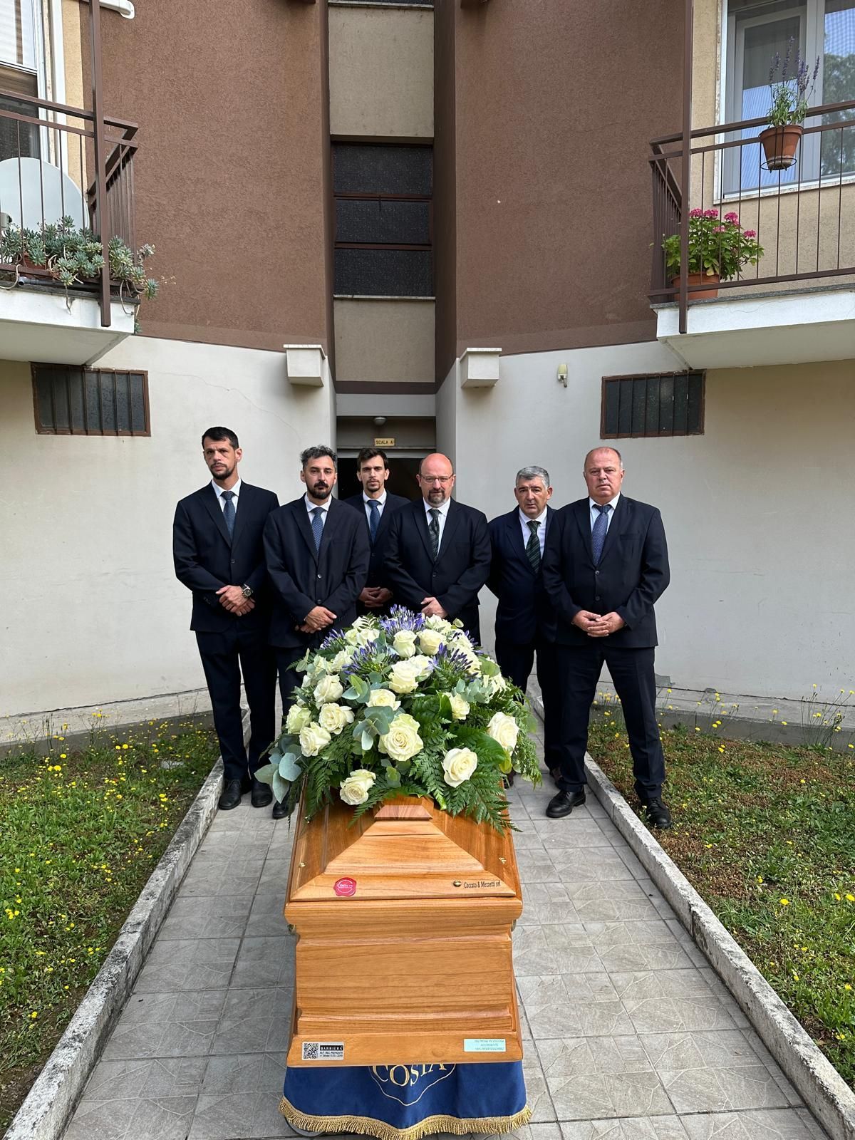 In un cortile, alcuni uomini in abiti scuri si trovano dietro una bara di legno ornata di fiori bianchi.