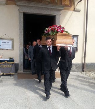 Uomini in abiti neri portano fuori dall'ingresso di un edificio una bara ricoperta di fiori.