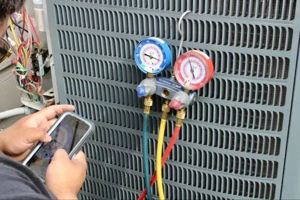 Technician checking HVAC refrigerant gauges on an outdoor unit with a phone in hand