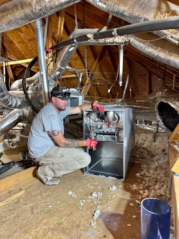 Worker kneeling in attic beside HVAC unit, installing ductwork and insulation.