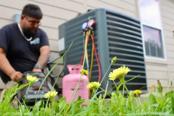 Technician working on outdoor HVAC unit beside pink propane tank and hoses in a grassy yard