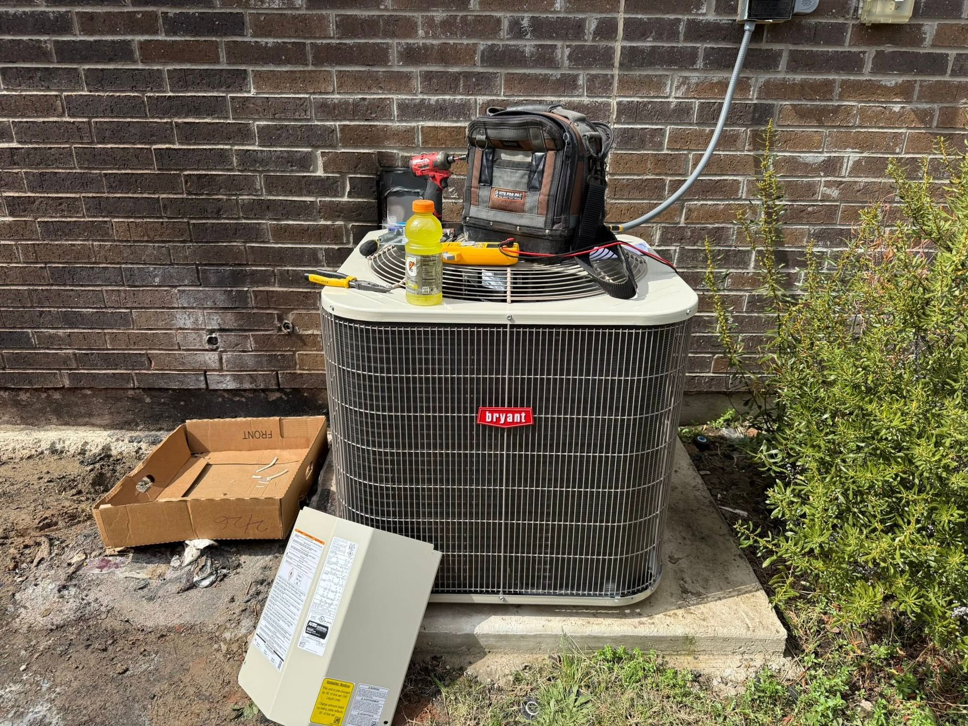 Air conditioner unit beside a brick wall with tools and supplies stacked on top, boxes on the ground
