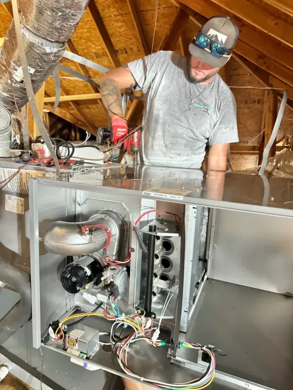 Worker in hard hat using a drill inside an exposed HVAC unit in an unfinished attic.