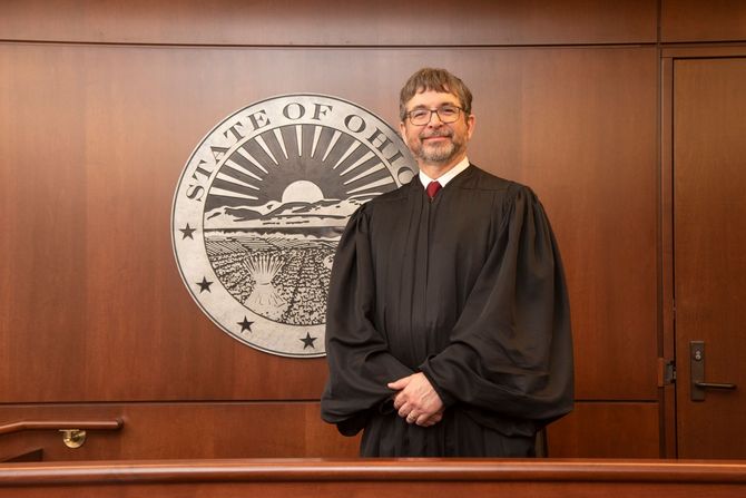 Judge Sylvain standing in a courtroom before the state seal, wearing a black robe and smiling.