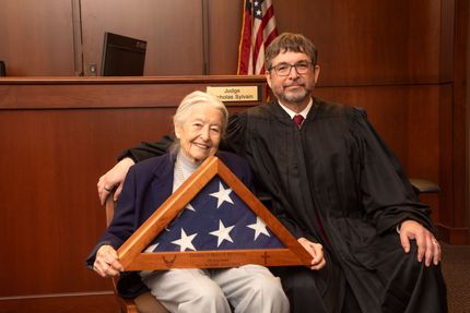 Judge Sylvain seated with his mom holding a folded American flag in a courtroom