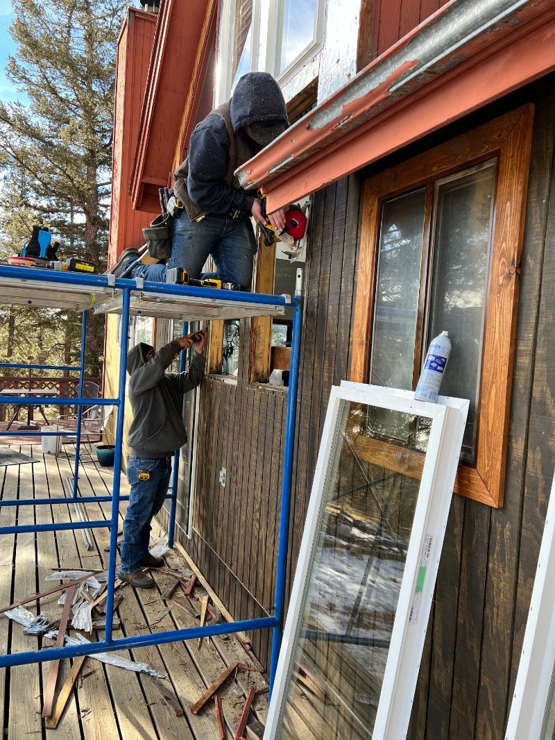 Two workers installing a window, one on a scaffold, the other at the building. Wooden siding on exterior.