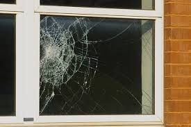 Broken window in a white frame on a brick building; glass shattered with spiderweb-like cracks.