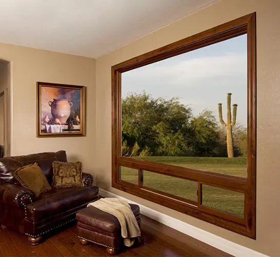 Living room with large wooden-framed window overlooking a landscape with a cactus. Leather furniture and painting on wall.