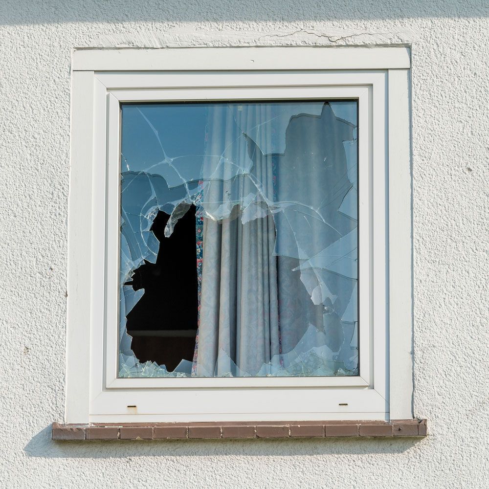 Broken window in a white frame, on a white textured wall, showing visible glass cracks and hole.