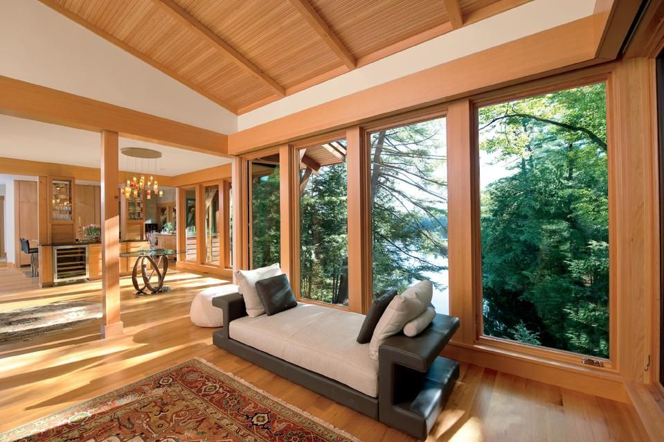 Bright, wooden-paneled room with large windows overlooking a lake and trees, featuring a chaise lounge and a patterned rug.