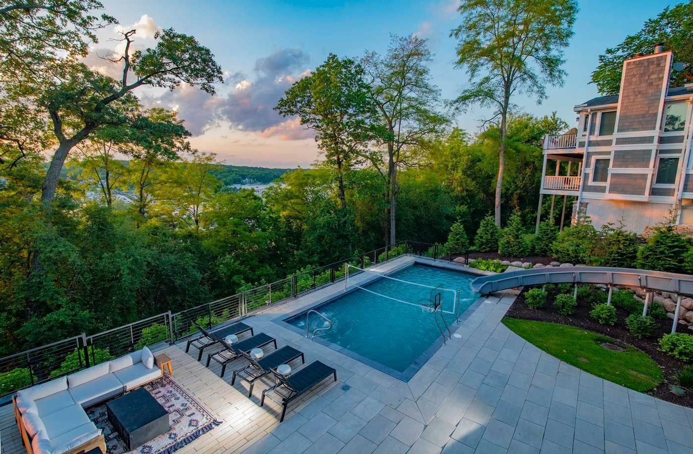 Backyard patio with a rectangular pool, lounge chairs, and wooded sunset views beside a modern house