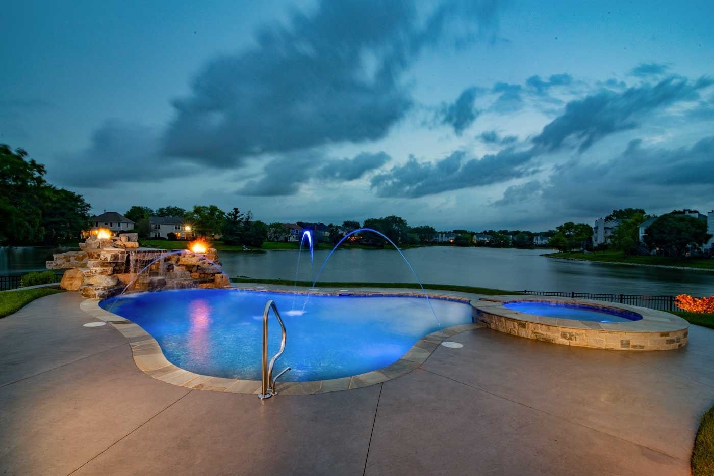 Twilight view of a lit outdoor pool beside a calm lake and stone patio