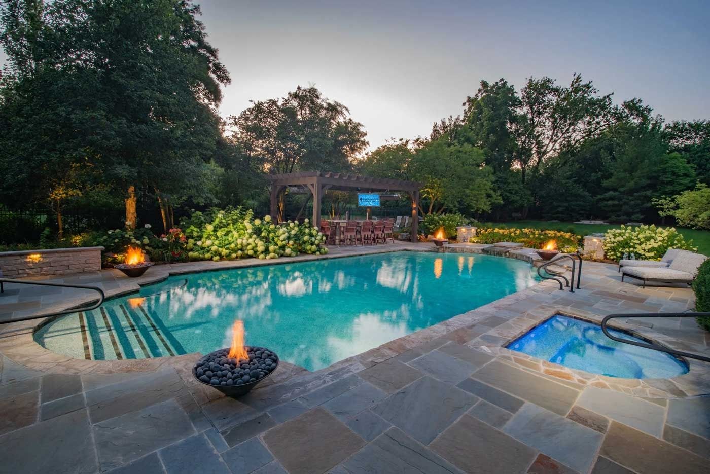Backyard pool with glowing fire bowls, spa, and lounge chairs at dusk