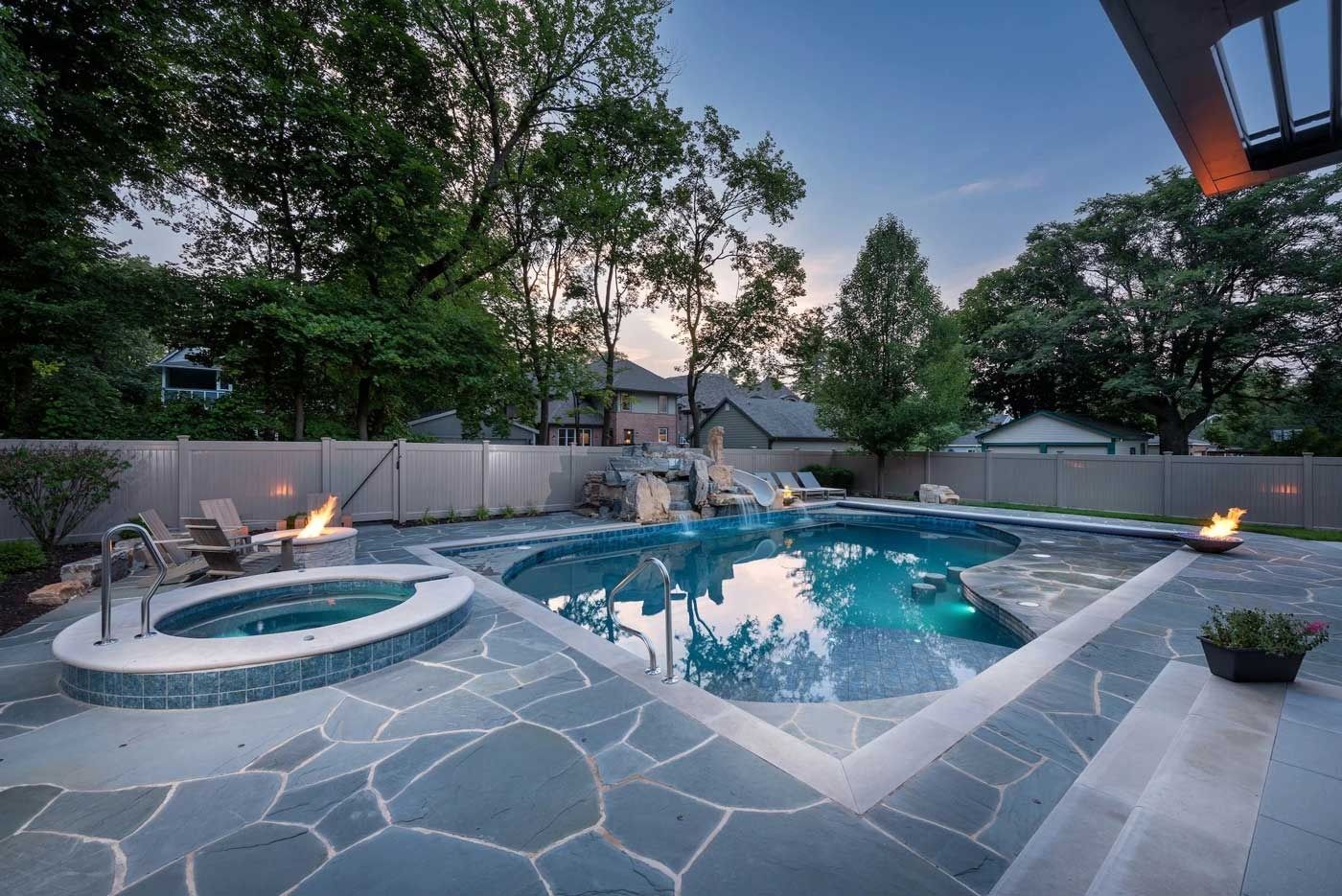 Backyard stone patio with a lit fire pit and a blue swimming pool at dusk