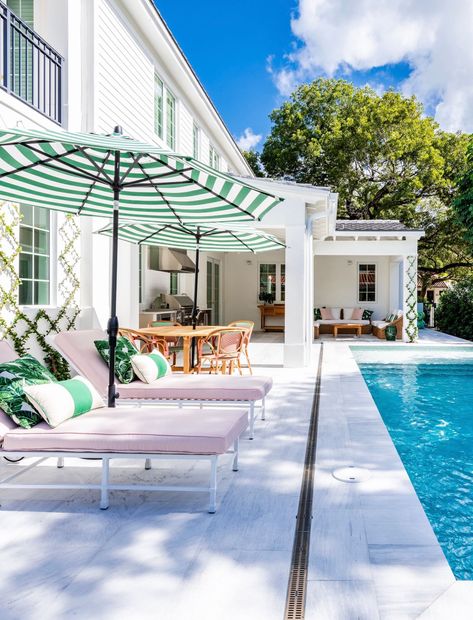 Sunny poolside patio with striped umbrellas, white lounge chairs, and a blue swimming pool beside a white house