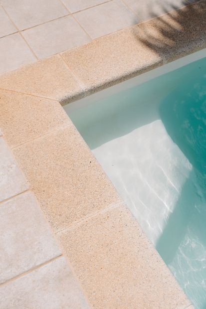 Corner of a turquoise swimming pool beside beige stone decking, with palm shadows on the pavement