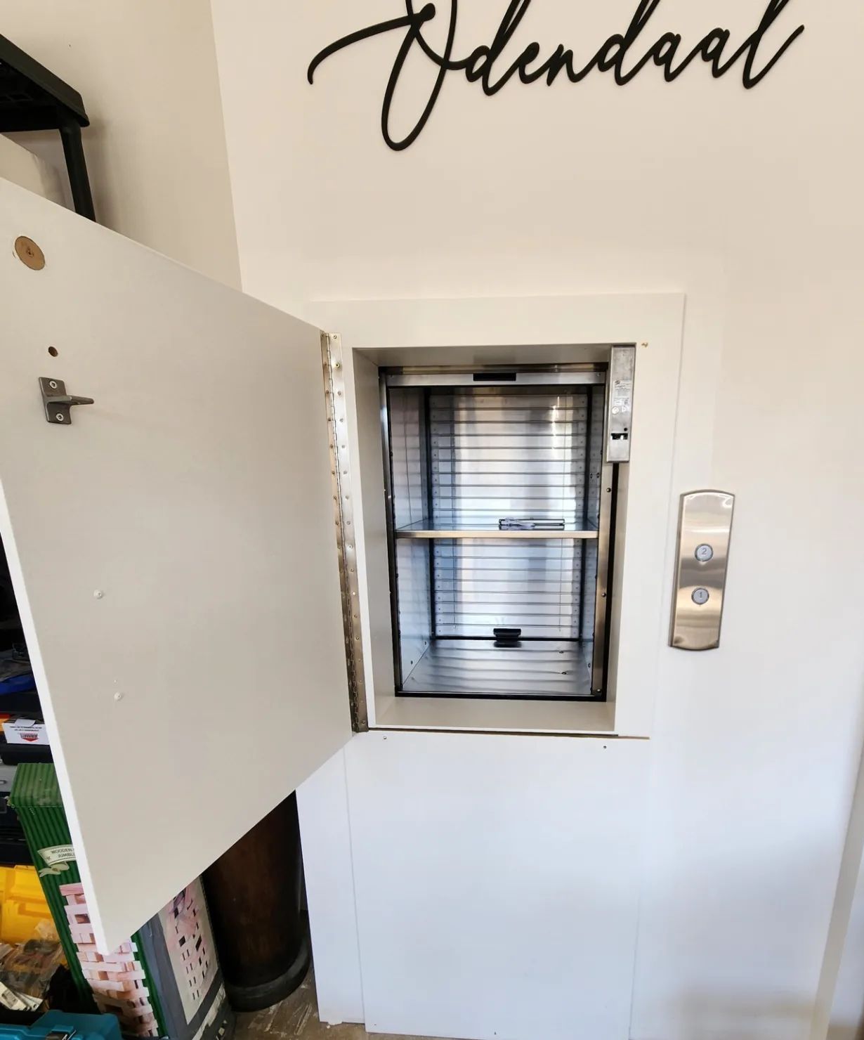Petit ascenseur mural avec une porte blanche ouverte dans un couloir, sous un panneau « Avendaal ».