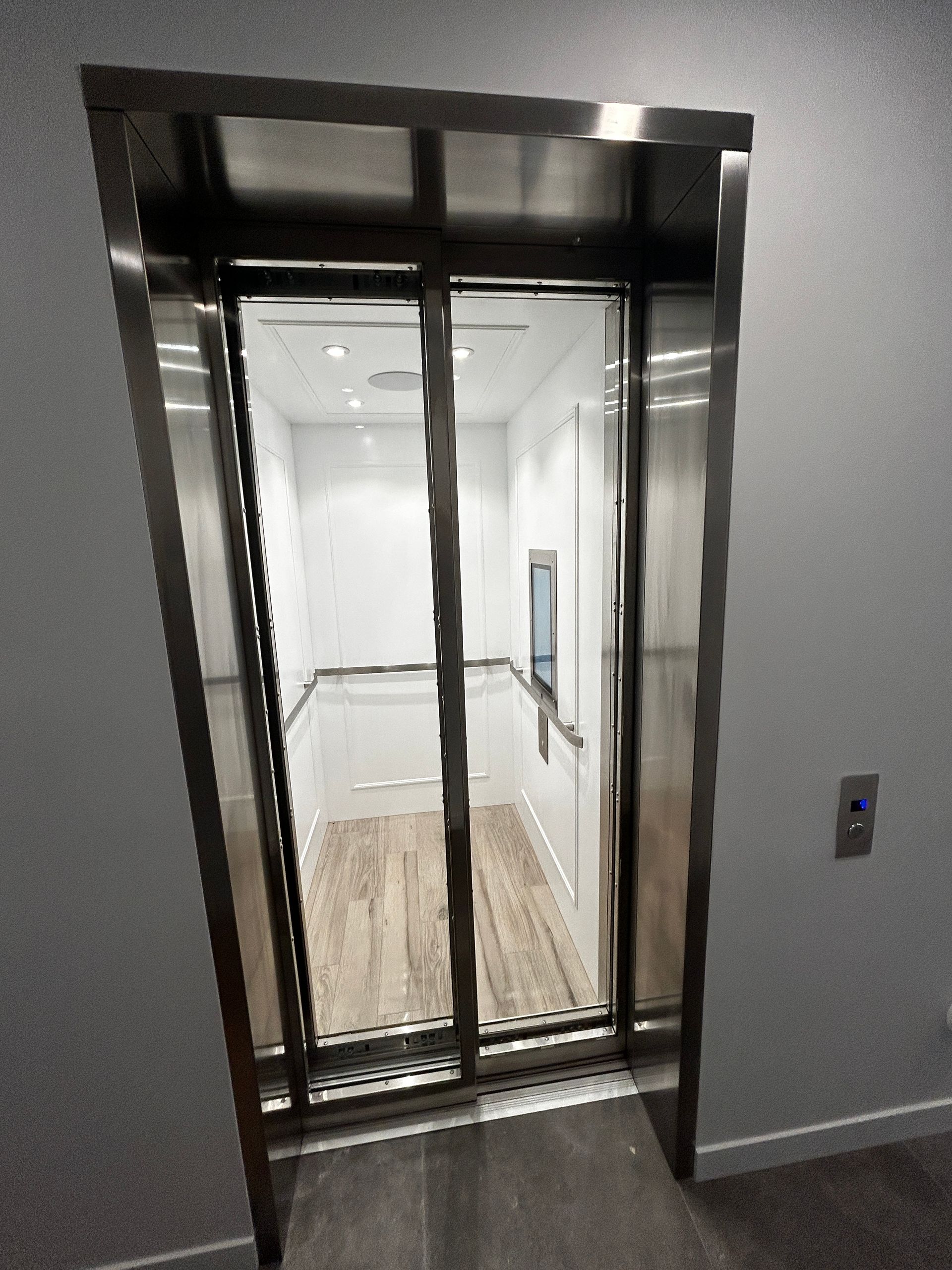 Ascenseur ouvert avec portes métalliques, intérieur blanc et sol imitation bois dans un couloir.