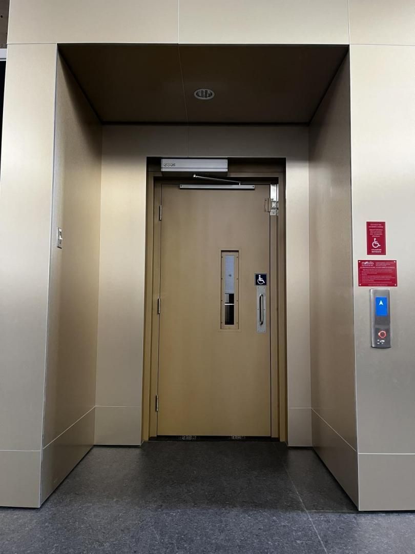Portes d'ascenseur beiges dans un couloir, avec un panneau d'alarme incendie rouge et un bouton d'appel sur la droite.