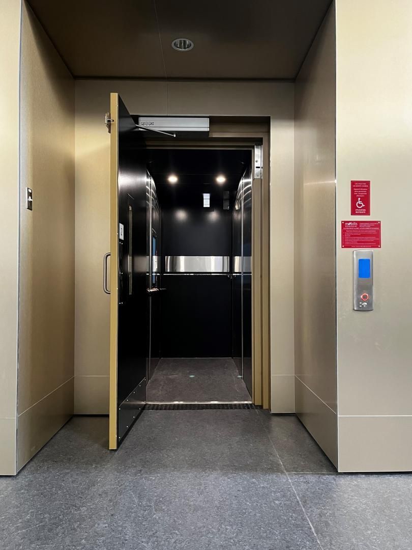 Ascenseur ouvert avec portes dorées dans un couloir beige, panneau de commande éclairé sur la droite.
