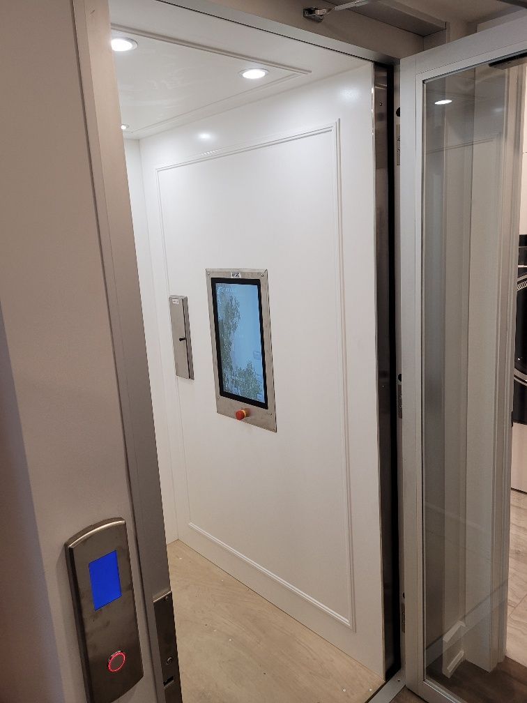 Ascenseur ouvert avec portes vitrées, intérieur blanc et panneau de commande mural dans un immeuble moderne.