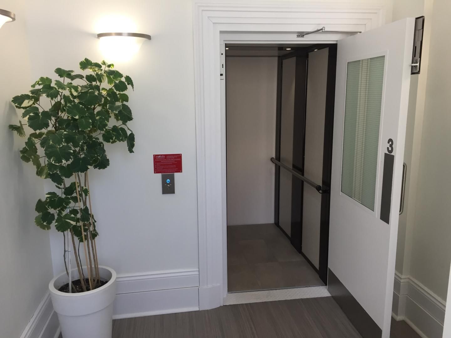 Hall d'ascenseur avec une plante en pot, des appliques murales et une porte d'ascenseur ouverte.