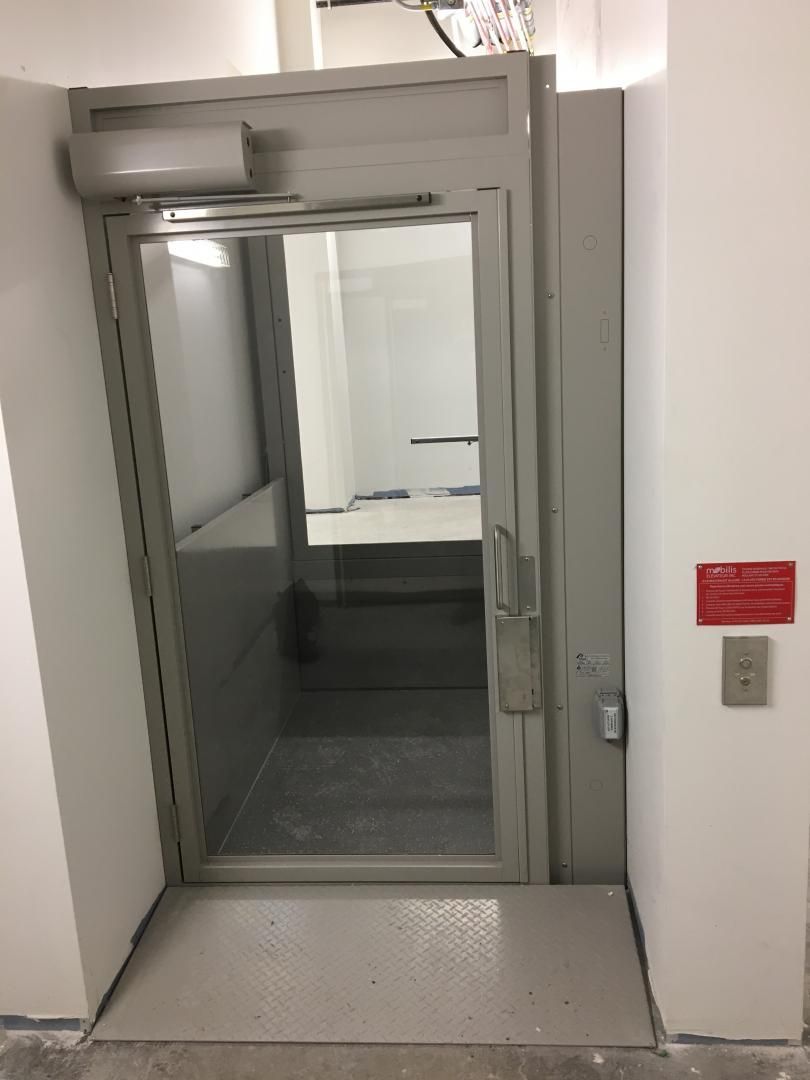 Porte accessible avec porte vitrée, rampe d'accès et bouton d'appel mural dans un couloir