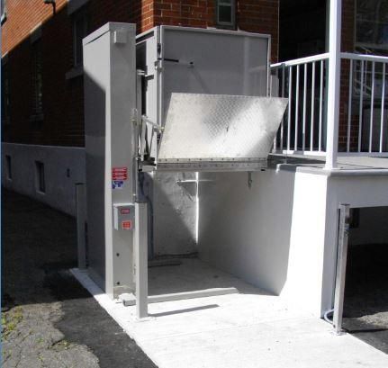 Plateforme élévatrice extérieure pour fauteuils roulants située à côté de l'entrée d'un bâtiment avec des garde-corps métalliques.
