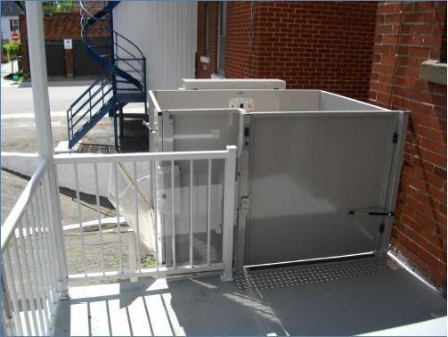 Plateforme élévatrice métallique blanche pour personnes à mobilité réduite, située à côté d'un bâtiment en briques et d'une rampe d'accès pour trottoir, avec barrière de sécurité fermée.