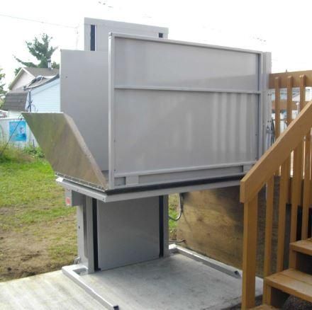 Plateforme élévatrice extérieure en métal avec boîtier gris, située à côté d'un escalier en bois sur une terrasse.