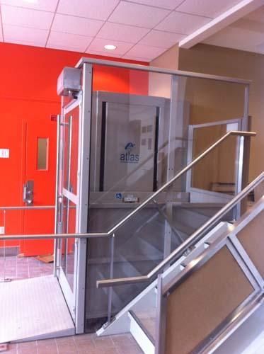 Ascenseur vitré pour personnes à mobilité réduite situé à côté d'un escalier dans un bâtiment aux murs rouges et aux rampes métalliques.