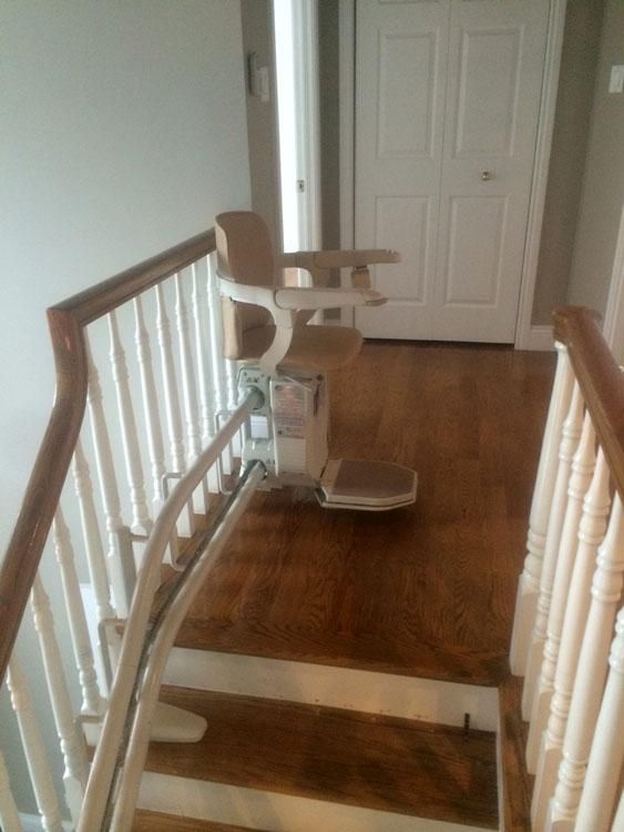 Un monte-escalier est installé sur un escalier en bois, à côté d'une rampe blanche, menant à une porte blanche fermée.