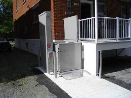 Ascenseur extérieur pour fauteuils roulants à côté d'un bâtiment en briques avec une rambarde blanche et une plateforme en béton avec rampe d'accès