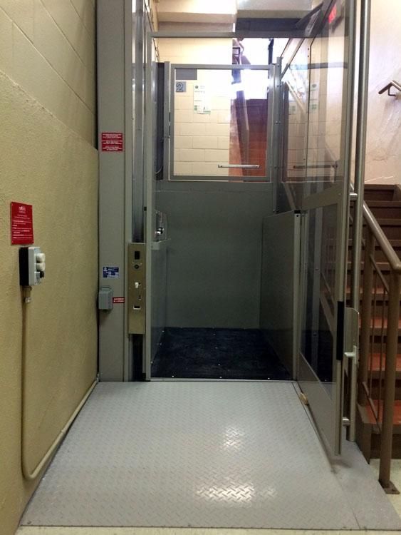 Couloir étroit avec un ascenseur vitré au fond et une rampe d'escalier métallique sur la droite.