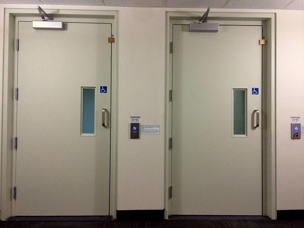 Deux portes de toilettes beiges avec des fenêtres étroites et des panneaux d'accessibilité bleus dans un couloir